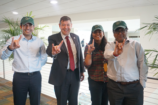 from left to right stand a young Indian male scholar, an older caucasian gentleman in a suit, a young female Indian scholar, and aother young male Indian scholar posing in USF green hats while proudly displaying the "Bulls" sign with their hands