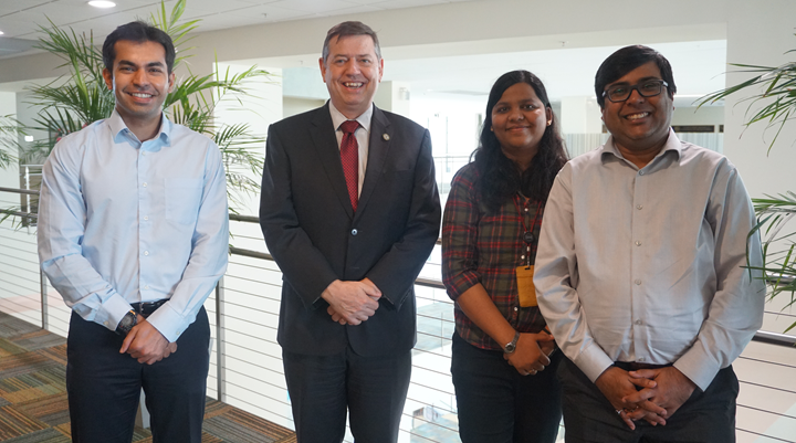 from left  to right stand a young Indian male scholar, an older caucasian gentleman in a suit, a young female Indian scholar, and aother young male Indian scholar posing professionally for the camera, 