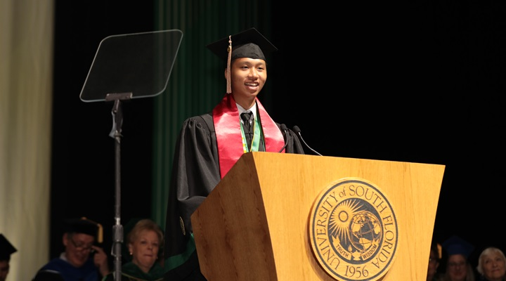 Vietnamese student, Kha Do stands at a podium on stage in regalia while giving a commencement speech