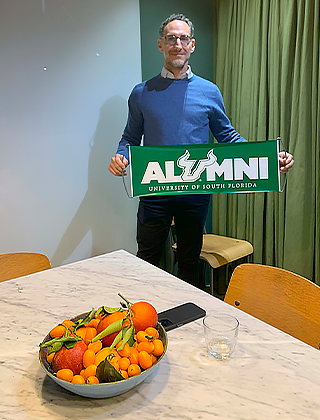 Arpesani holding a green USF sign that says "Alumni" and in the foreground is a pot of fruit