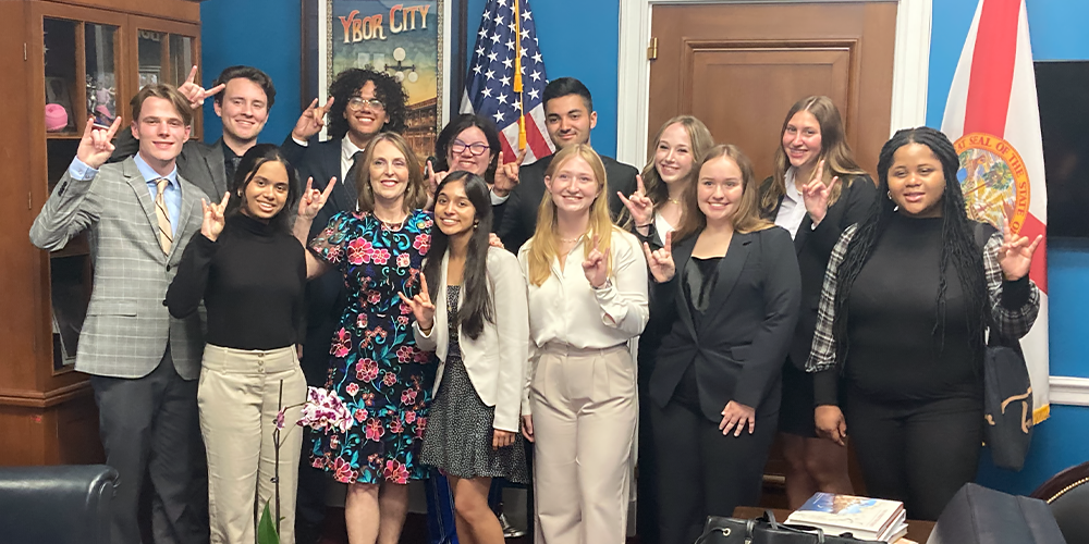 A group of students holding up the "bulls" sign and standing in an official room with the U.S. and Florida flag in the background