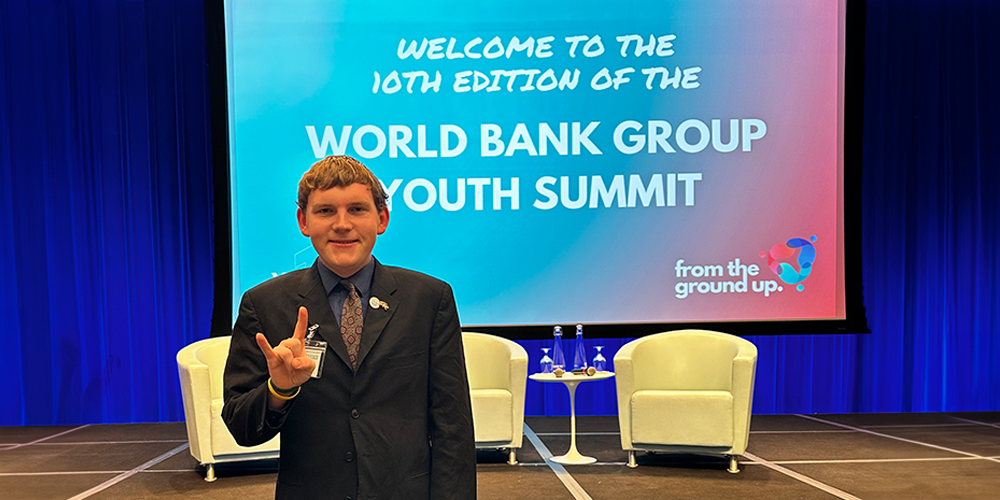 young white male standing in a dark suit on a stage infront of the welcome to the 10th edition of the World Bank Group Summit welcome sign