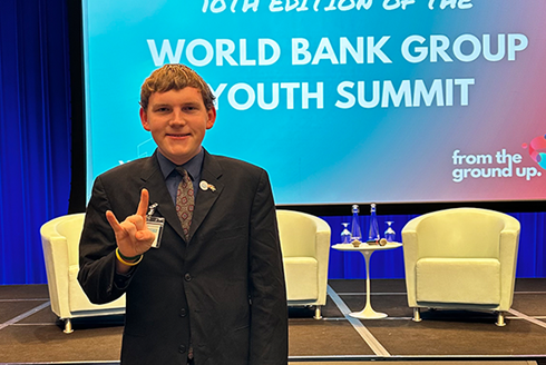 young white male standing in a dark suit on a stage infront of the welcome to the 10th edition of the World Bank Group Summit welcome sign