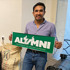 USF global alumnus, Hernan Herrera poses with an alumni sign amongst his coffee import and roasting business