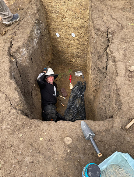 Student wearing a safety hat laying in a grave with excavation equipment