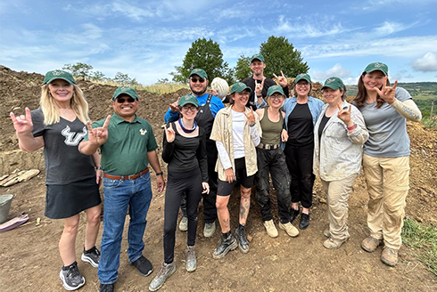 Students, Bethard, Dr. Kiki Caruson, and Provost Mohapatra stand at Transylvania burial site raising their hands with the bull sign