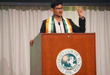 Student body president speaks at podium and raises the "bulls" sign with his hand