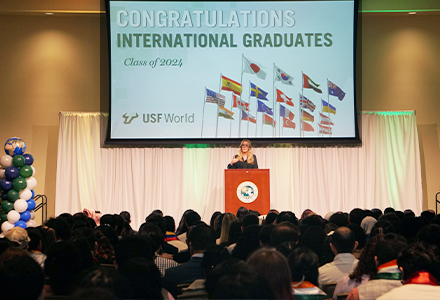 Dr. Kiki Caruson speaks at a podium in front of a large room filled with international students and families