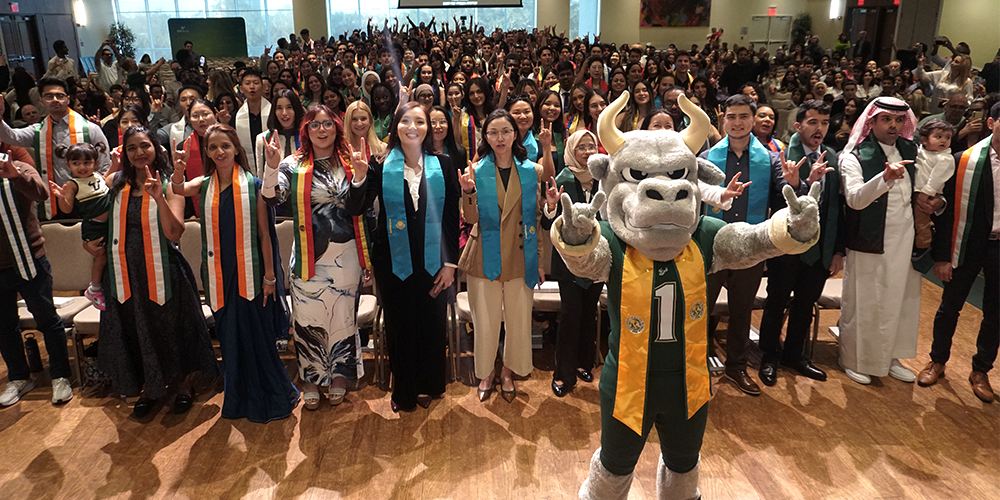 A group of students with different colored sashes raising their hands with bull sign, Rocky the Bull USF mascot is standing in front of the group holding up the bull sign