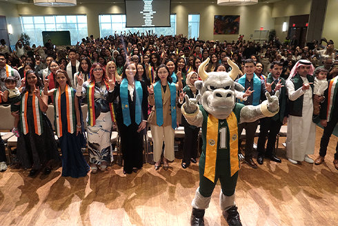 A group of students with different colored sashes raising their hands with bull sign, Rocky the Bull USF mascot is standing in front of the group holding up the bull sign