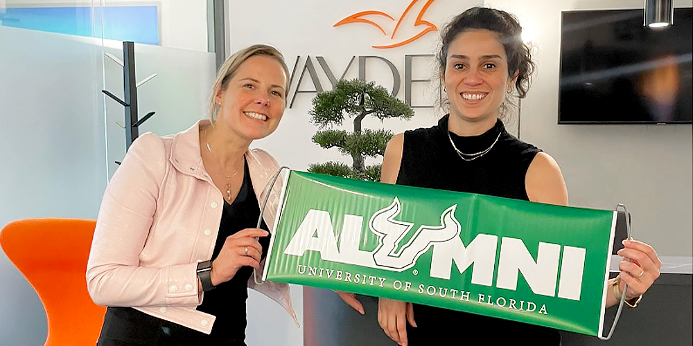 Alumnus Alexandra posing with Vanessa Martinez and both holding the green Bulls Alumni Banner