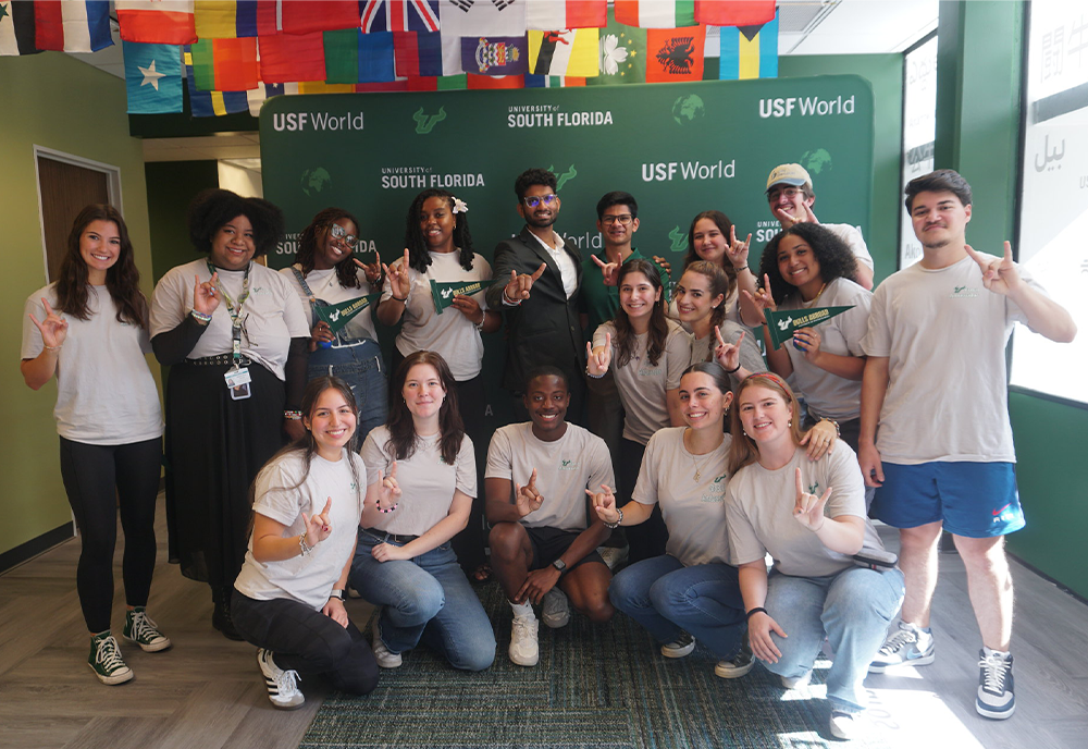 Students giving the bulls sign in front of USF World green backdrop and international flags hanging above them from the ceiling