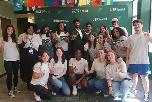 Students giving the bulls sign in front of USF World green backdrop and international flags hanging above them from the ceiling