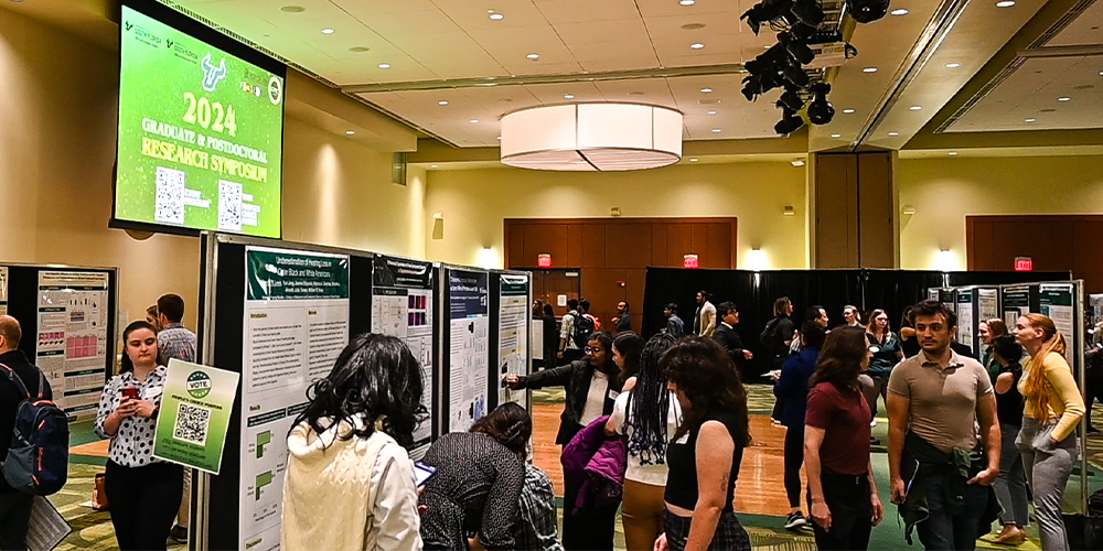 room view of scholars standing with their research posters and speaking with symposium guests