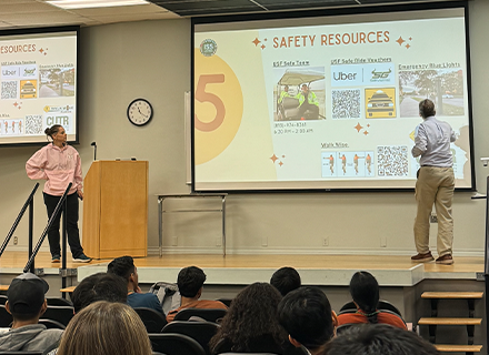 An auditorium full of people watching a presenation given by two people on stage