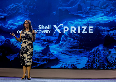 Virmani giving a speech in front of a screen with a 3D oceanongraph and the words "Shell Oceans Discovery X Prize"