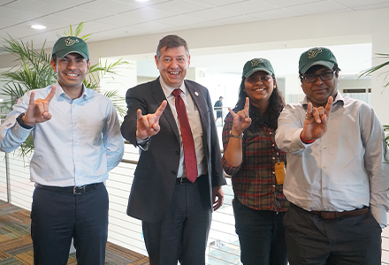 from left to right stand a young Indian male scholar, an older caucasian gentleman in a suit, a young female Indian scholar, and aother young male Indian scholar posing in USF green hats while proudly displaying the bulls sign with their hands