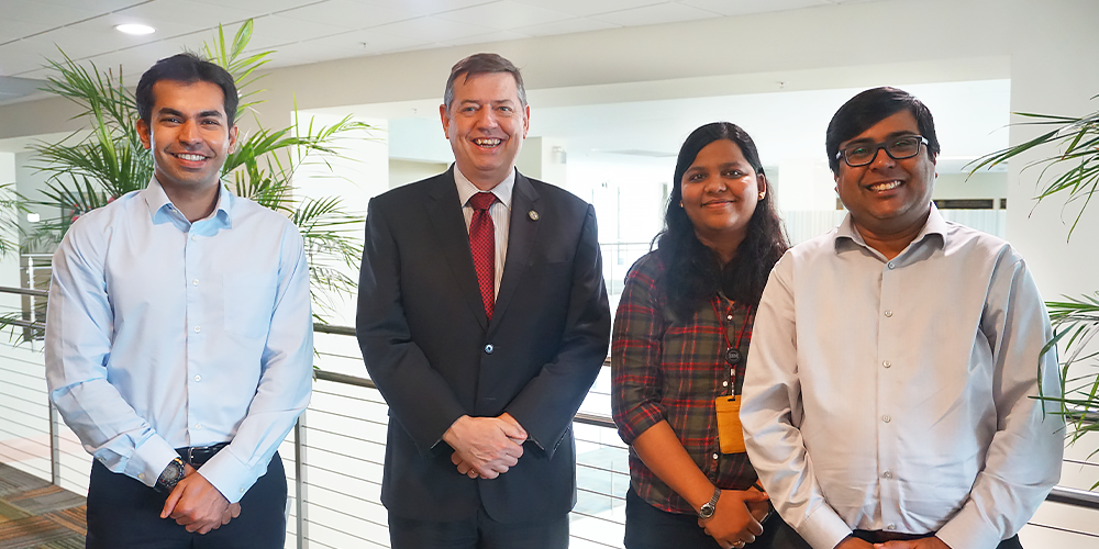 from left  to right stand a young Indian male scholar, an older Caucasian gentleman in a suit, a young female Indian scholar, and another young male Indian scholar posing professionally for the camera