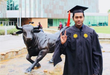 khan with graduation cap and gown on making the bulls sign with his hand in front of the bulls statue on USF campus