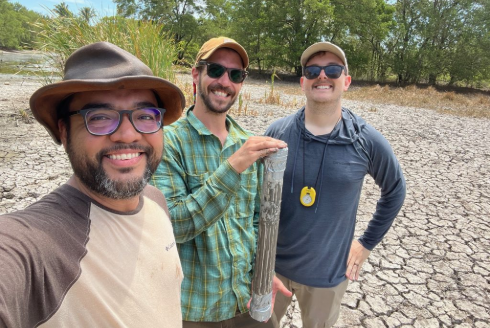 Three men smile at the camera while standing in a forested area with cracked earth under their feet and green tress in the background