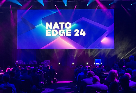 A large dark room filled with people and a large screen that says "Nato Edge 24"