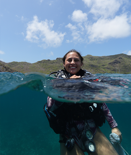 Tatiana in the ocean in scuba gear, smiling at camera which is half in water and half above