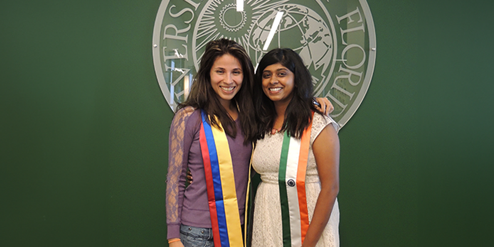 Neha standing beside another person, both wearing sashes with the flag colors of their home countries